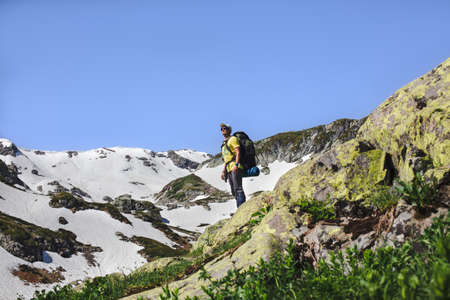 Young male conqueror of the summit wearing climbing clothing and a backpack, looks at horizon with a...