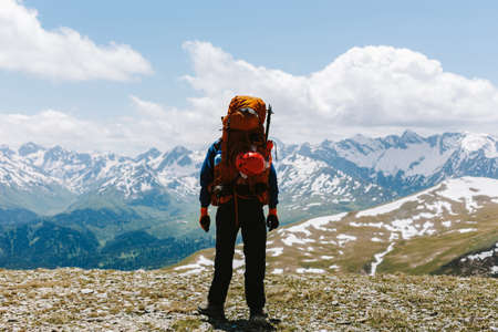 Rear view of a man tourist mountaineer in sportswear and large backpack stands on top of mountain and admires picturesque view of valley and mountain ranges covered with snow with white cloudsの写真素材