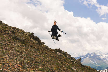 Happy young female tourist jumps on top of a mountain with climbing sticks in her hands against the...