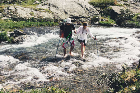 Tourists with ski poles cross the stormy mountain stream while traveling in the mountains on a warm...
