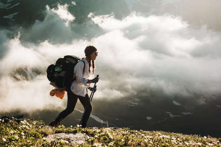 Side view of young tourist girl with large backpack walking along top of mountain with wildflowers on background of charming view of white clouds and snow-capped mountains. Concept of mountaineeringの写真素材