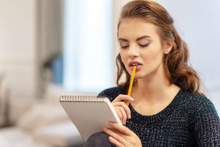 Ideas for business. Studying and working at home. Thoughtful young woman making notes using notepad in kitchen.の写真素材