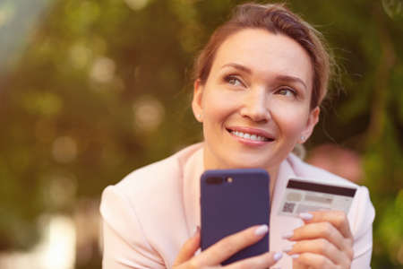 Businesswoman with a credit card and phone makes purchasing outdoors. Successful woman is using smart phone and bank card for online shopping.の写真素材