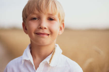 Face close - up of a boy of eight years. Portrait of a smiling boy in nature looking at the camera. Happy blond boy with a smile on his face. Child in a white shirt with blue eyes in nature. の写真素材