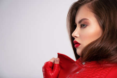 Profile portrait of beautiful young woman with bright makeup. Beautiful brunette with bright red lipstick on her lips. Pretty girl with long brown hair. Brunette in a black leather jacket.の写真素材