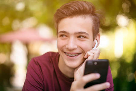 Happy teenage boy is using mobile phone, outdoors.  Close-up portrait of a smiling young man with smartphone, in park.  Cheerful teenager in casual clothes with cell phone in park. Soft focusの写真素材