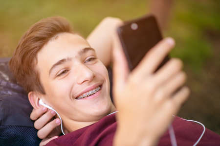 Happy teenage boy is using mobile phone, outdoors.  Close-up portrait of a smiling young man with...