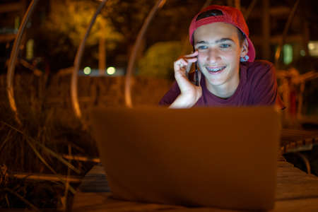 Portrait of a smiling young man with a smartphone, in the street at night.  Happy teenage boy is...
