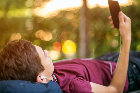 Teen boy with smartphone lies on the bench in the park.   Teenage boy is using phone, outdoors. ...