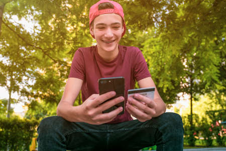 Teenage boy with a credit card and phone makes purchasing outdoors. Happy young man is using smart phone and bank card for online shopping. Handsome smiling white guy holds bank  card and cell phoneの写真素材