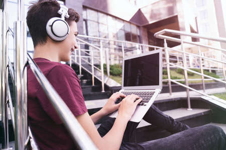 Smiling teenage boy with a laptop on the street. Handsome young man works on a notebook, outdoors....