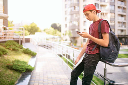 Young man  stands with backpack and holds smartphone, in the city.   Teenage boy is using mobile phone, outdoors.  Caucasian teenager in casual clothes with cell phone, urban scene.の写真素材