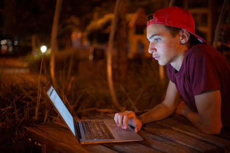 Teenage boy with a laptop in the park. Handsome young man works on a notebook, outdoors. Caucasian guy holds a laptop on the knees and looking to the screen. Teenager in headphones with laptopの写真素材