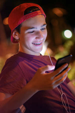 Close-up portrait of a smiling young man with a smartphone, in the street.  Happy teenage boy is using mobile phone, outdoors. Cheerful teenager spends time in social networks using cell phone.の写真素材