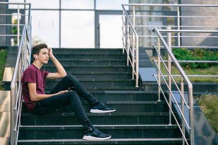 Dreaming guy in earphones holds a mobile phone in hands.  Relaxing teenage boy with calm emotions on his face look into the distance. Handsome young man listens to the music, outdoors. Happiness.の写真素材