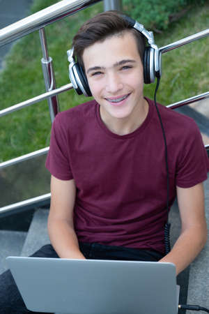Smiling teenage boy with a laptop on the street. Handsome young man works on a notebook, outdoors. Cheerful guy holds a laptop on the knees and looking to the screen. Teenager in headphones with laptopの写真素材