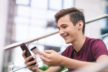 Teenage boy with a credit card and mobile phone makes purchasing outdoors. Happy young man is using...