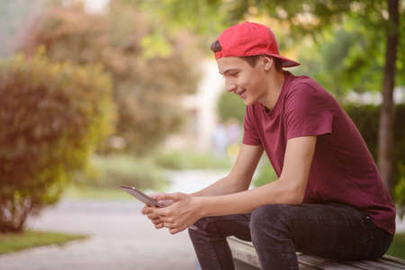 Teenager spends time in social networks. Smiling teenage boy with a tablet in the park. Handsome young man works on a tablet outdoors. Cheerful guy holds a tablet on the knees and looking to screen.の写真素材