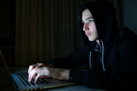 Hacker is  hacking into the computer network. Young boy in a dark room with laptop.  Hacker with a...