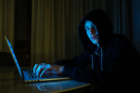 Hacker is  hacking into the computer network. Young boy in a dark room with laptop.  Hacker with a...
