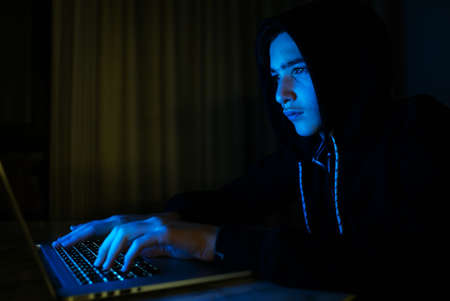Hacker is  hacking into the computer network. Young boy in a dark room with laptop.  Hacker with a...