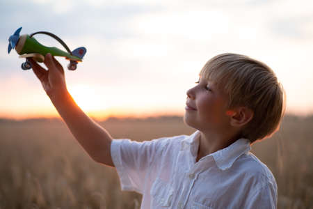 Happy child playing with a toy plane in nature during summer sunset. Boy in a  white shirt with a plane in hands on wheat field. Kid holds a wooden airplane and dreams of being a pilot, on the natureの写真素材