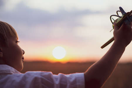 Boy in a  white shirt with a plane in hands against sky. Kid holds a wooden airplane and dreams of being a pilot, on the nature. Happy child playing with a toy plane in nature during summer sunset.の写真素材