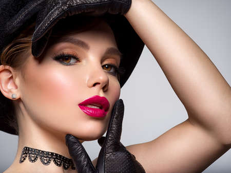 Art portrait of beautiful young woman with orange makeup in a black hat.  Closeup face of a sexy girl with red lipstick on lips. Stylish fashion portrait. Attractive model with a black gloves at studio.の写真素材