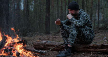 Man with cup of tea the fire in the forest. Human looks at the bonfire and drinks tea on the nature in the summer. Alone traveler sits and rests by the fire with a cup of coffee at dusk.の写真素材