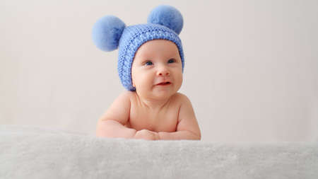 A smiling infant baby in a blue hat with pompoms looks into the camera. A happy newborn is lying on a blanket. Beautiful, happy little child with blue eyes.  Cheerful White kid.の写真素材