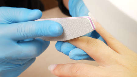 Manicurist files the shape of the nails with a special nail file, close up view. The manicurist shapes the nails with a nail file. The process of nail care in a beauty salon. Nail file processing.の写真素材