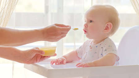 Mom feeds a 9 month baby with fruit puree from a spoon, close-up, high key.  Mother is Feeding a baby from a spoon.  Infant boy eats sitting on baby's chair. Mother cares about little son.の写真素材