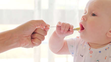 Mother is Feeding a baby from a spoon. Mom feeds a 9 month baby with fruit puree from a spoon, close-up, high key. Infant boy eats sitting on baby's chair. Mother cares about little son.の写真素材