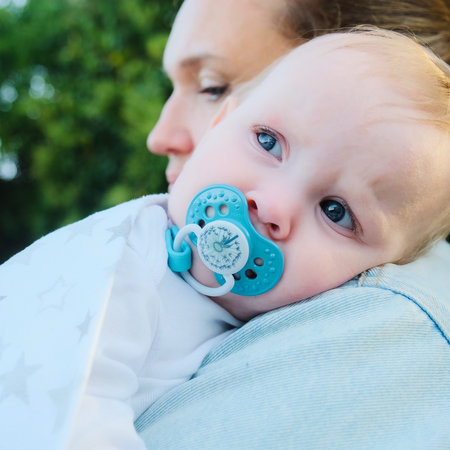 Baby rests on his mother's shoulder, in park. Close-up of a calm and looking away little child on...