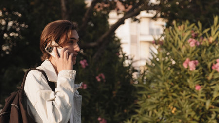 Young man is talking on a cell phone while walking down the street. Positive guy is talking on the phone on the street. Handsome man in the park communicates using a mobile phone. Communication.の写真素材