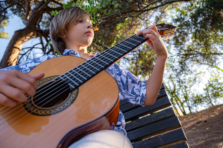 Boy plays the guitar in the park sitting on a bench.  Child plays a musical instrument, in nature.  Guitarist plays on guitar outdoors.  Musician plays a classical guitar in the park. Hobby concept.の写真素材