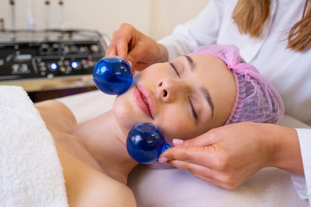 Beautician making a massage of the  face using the blue balls. Woman in a spa salon on cosmetic procedures for facial care. Girl gets massage of a face. Cosmetologist makes face massage.の写真素材