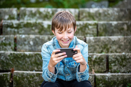 A happy young kid is sitting outside with a phone. Smiling teen boy outdoors looking at the...