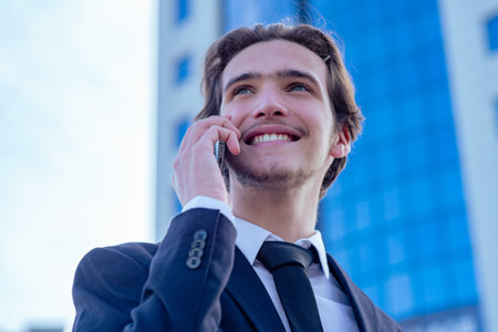 Smiling businessman near a business center calling by cell phone. A happy young businessman talking with a smartphone. Young cheerful businessman near a modern tall glass building with a phone.の写真素材