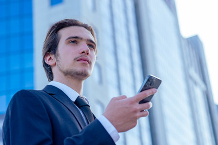 Confident businessman near a business center with a cell phone. A young businessman in a suit is looking at a smartphone. A young businessman near a modern tall glass building with a mobile phone.の写真素材