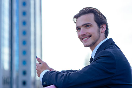 Smiling businessman near a business center with a cell phone. A happy young businessman looking at a smartphone. A young cheerful businessman near a modern tall glass building with a mobile phone.の写真素材
