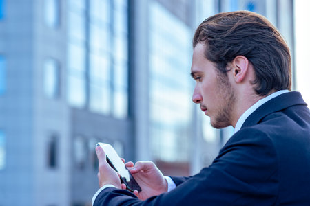 Confident businessman near a business center with a cell phone. A young businessman in a suit is looking at a smartphone. A young businessman near a modern tall glass building with a mobile phone.の写真素材