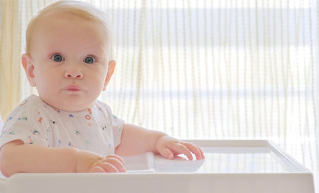 Cute baby is sitting on a high chair waiting for a meal. Beautiful white-haired toddler is sitting in a high children's chair at home.  Cute blond little boy looks at the camera.の写真素材