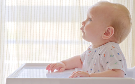 Cute baby is sitting on a high chair waiting for a meal. Beautiful white-haired toddler is sitting in a high children's chair at home.の写真素材