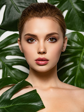 Beautiful woman with green leave near face and body. Closeup girl's face with green leave. Pretty girl in Skin care beauty treatments concept. Model with big tropical leaf, posing, studio. Brown makeup.の写真素材