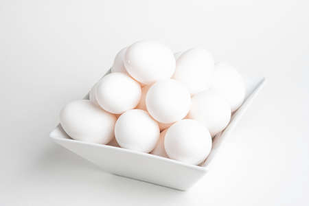 Fresh white eggs piled atop a square white porcelain bowl and set on a white background.