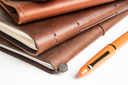 A stack of leather-bound travel writing journals with an orange fountain pen set on plain white background,の写真素材