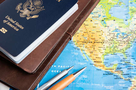 The foil-stamped dark blue front cover of an American passport with leather-covered travel notebooks and writing pen set on a world map background.の写真素材