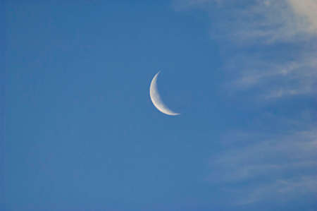 A slim sliver of silvery moon set on an expanse of a partly cloudy blue sky.