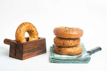 A single tall stack of three freshly baked bagels on a napkin with one bagel on a cutting wood stand and knife set on a plain white background.の写真素材